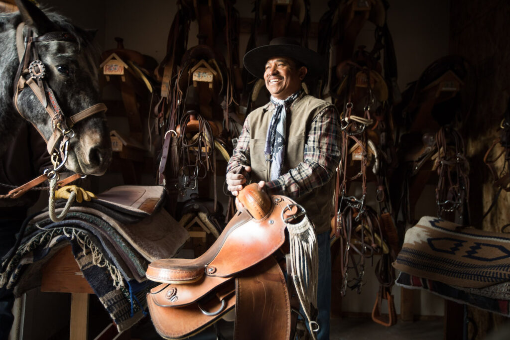 Man smiling in horse stables while holding a saddle